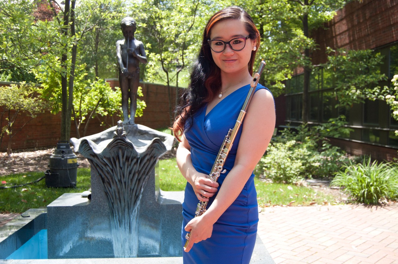 A graduate student poses with her flute in front of a fountain on campus.